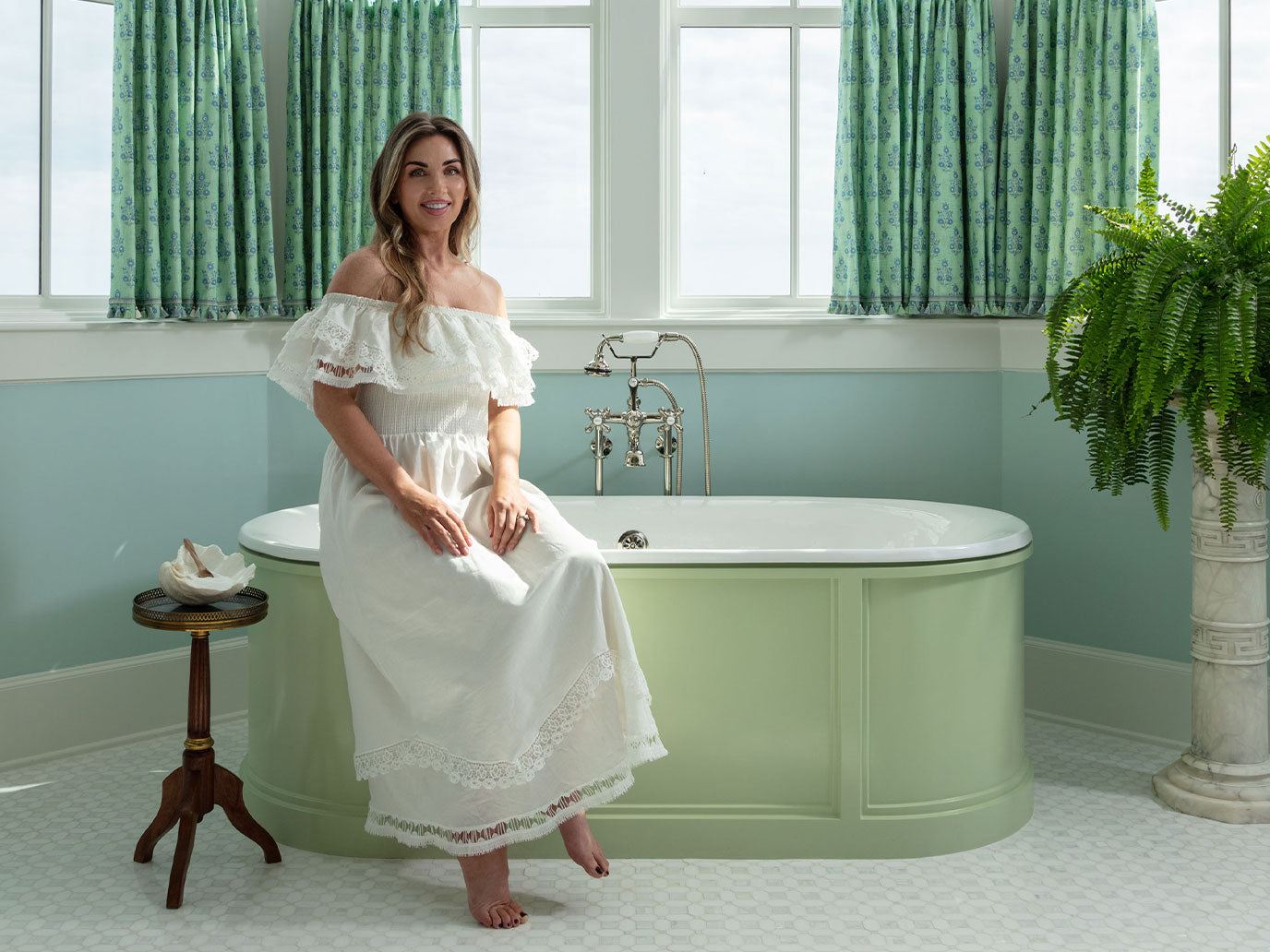Woman in a white dress sitting by a green bathtub in a bathroom with green curtains and a plant.